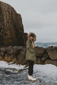 a woman in a green parka standing on a rock next to the ocean